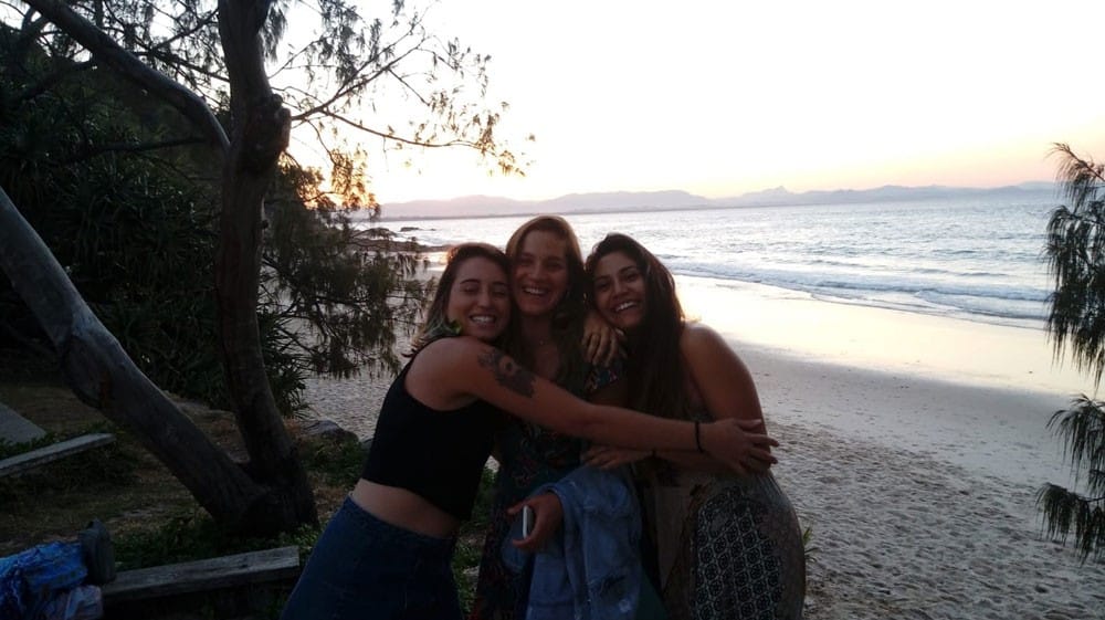 grupo de 3 chicas sonrientes mirando a cámara y detrás la playa y la puesta de sol